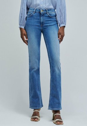 Jeans a zampa d'elefante blu in un lavaggio medio con vita alta, chiusura con bottone e cuciture visibili. Completati da sandali con tacco bianchi.