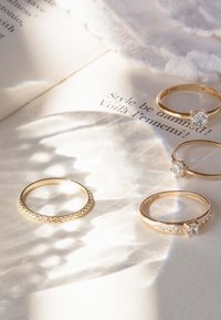 Quatre bagues en or disposées sur un fond clair, comprenant une bande tressée, deux avec des pierres claires, et une avec un petit accent en diamant.