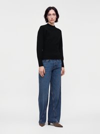 Jeune femme aux cheveux courts et foncés, portant un haut noir à manches longues, un jean bleu à jambes larges et des chaussures marron pointues, debout sur fond blanc.