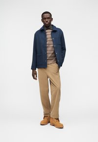 Μπλε μάλλινο σακάκι με λεπτομέρειες κουμπιών, μπεζ παντελόνι από κορδόνι, πουλόβερ με σχέδια και ταμπά μποτάκια εργασίας. Το μοντέλο στέκεται μπροστά σε λευκό φόντο.