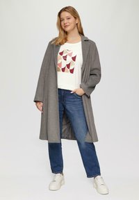 Grijze lange jas met brede mouwen over een wit T-shirt met een geometrisch patroon in rood, roze en goud, gecombineerd met blauwe jeans en witte sneakers.