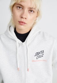 Haut de sweat à capuche zippé léger gris clair en tissu doux, avec un texte noir "SANTA CRUZ" et une broderie orange "Skateboards". Détails avec cordon de serrage.