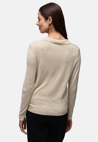 Femme aux longs cheveux bruns portant une chemise beige clair à manches longues et un pantalon noir, vue de dos sur un fond uni.