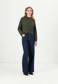 Pullover accorciato verde oliva con texture a coste, abbinato a jeans a gamba larga blu scuro con dettagli delle tasche laterali e cuciture minime.