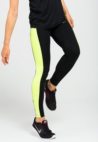 Leggings neri con un pannello laterale giallo neon, design aderente, texture morbida, abbinati a scarpe sportive nere con accenti rosa.