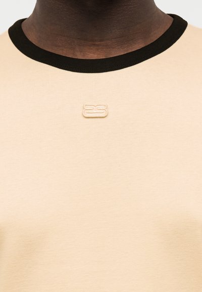 Μπεζ μπλουζάκι με μαύρο στρογγυλό λαιμόκοψη που φοριέται από ένα άτομο, με μικρό μπεζ κεντημένο λογότυπο "2S" τοποθετημένο κοντά στο γιακά.