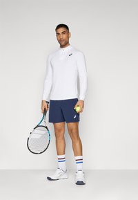 Hvid langærmet træningsshirt med krave, marineblå shorts og hvide tennissko. Holder en tennisketsjer og en grøn tenniskugle.