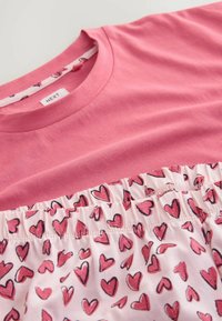 Růžové bavlněné tričko s nařaseným lemem a vzorem srdce ve světlé a tmavě růžové na bílém pozadí. Uvnitř výstřihu je viditelná visačka.