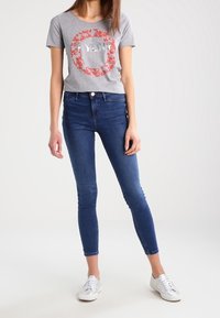 Grå t-shirt med en röd blommig cirkeldiagram och text, ihop med mörkblå skinny jeans och vita sneakers. Enkel design och passform.