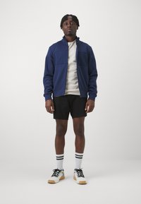 Marinefarvet zip-up jakke med to forlommer, grå T-shirt, sorte shorts, hvide sokker med sorte striber og hvide sneakers med gummisåler.