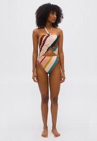 Γυναίκα με σγουρά μαλλιά, που φοράει ολόσωμο μαγιό σε ρίγες με πολύχρωμα χρώματα και με λαιμόκοψη halter και άνοιγμα μπροστά, όρθια ξυπόλυτη μπροστά σε απλό φόντο.