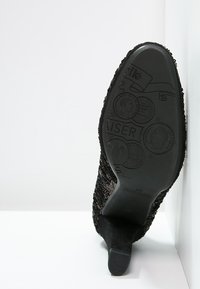 Semelle de chaussure noire avec surface texturée, présentant un talon arrondi et des motifs circulaires en relief. Le matériau semble en caoutchouc avec des détails complexes.