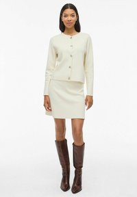 Cardigan en tricot ivoire avec col rond et fermeture à boutons, associé à une jupe ajustée assortie. Bottes en cuir marron jusqu'aux genoux.