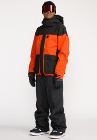 Veste de snowboard orange et noire ; dotée d'une capuche, de poches zippées et d'une coupe décontractée avec des poignets élastiques et un ourlet ajustable.