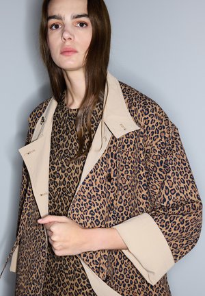 Jeune femme aux longs cheveux bruns portant une robe et un manteau assortis à imprimé léopard avec un col et des poignets beige, debout devant un fond gris uni.