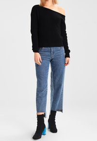 Femme portant un pull noir à épaules dénudées, un jean bleu court aux ourlets effilochés, et des bottines noires à talons carrés bleu vif.