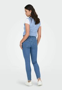 Chaleco de punto azul claro con patrones hexagonales, llevado sobre una camiseta blanca. Jeans azules ajustados con dobladillo desflejado y bolsillos traseros.