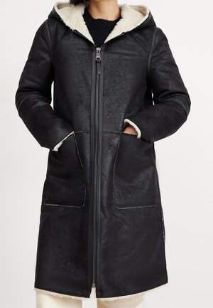 Veste longue en peau lainée noire avec fermeture éclair à l'avant, capuche, grandes poches avant et doublure en fourrure blanche visible à la capuche et aux poignets.