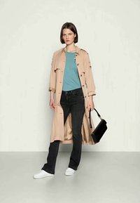 Casaco trench bege com botões, t-shirt teal, jeans escuros e ténis brancos. A segurar uma bolsa preta com um acabamento brilhante.