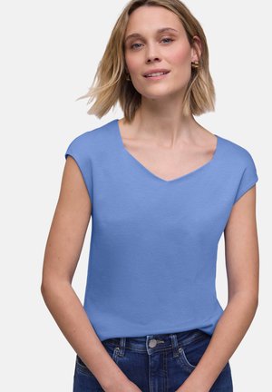 Frau mit kurzen blonden Haaren, die goldene Creolen, ein ärmelloses hellblaues Top mit V-Ausschnitt und dunkelblaue Jeans vor weißem Hintergrund trägt.