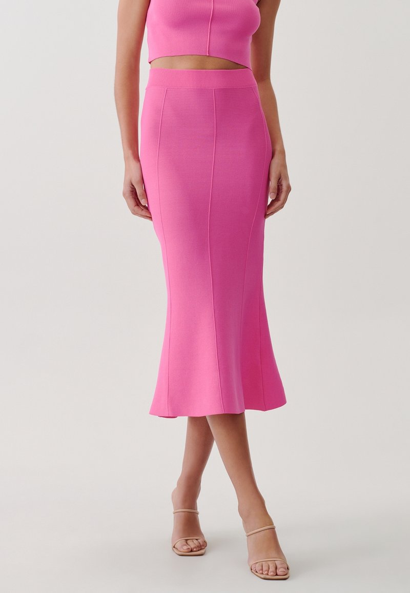 Femme portant une jupe midi rose ajustée et un top court assorti, associée à des sandales à talons couleur nude, debout jambes croisées.