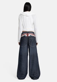 Ženska z dolgimi črnimi lasmi nosi bel pulover s kapuco in široke temne kavbojke z visokim pasom z zastavo Združenega kraljestva, obrnjena je stran.