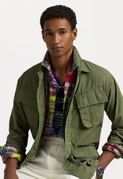 Casaco verde-oliva com fecho de botões e bolsos de lapela, sobrepõe-se a uma camisa estampada multicolorida com vários desenhos e riscas.