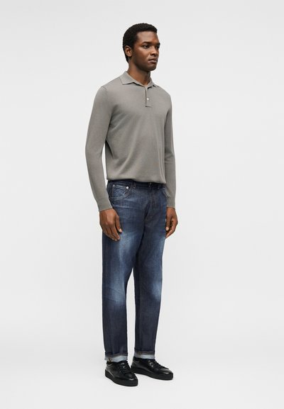 Άνδρας στέκεται φορώντας γκρι μακρυμάνικη πόλο μπλούζα, μπλε τζιν με γυρισμένα μπατζάκια και μαύρα αθλητικά παπούτσια σε απλό ανοιχτό φόντο.