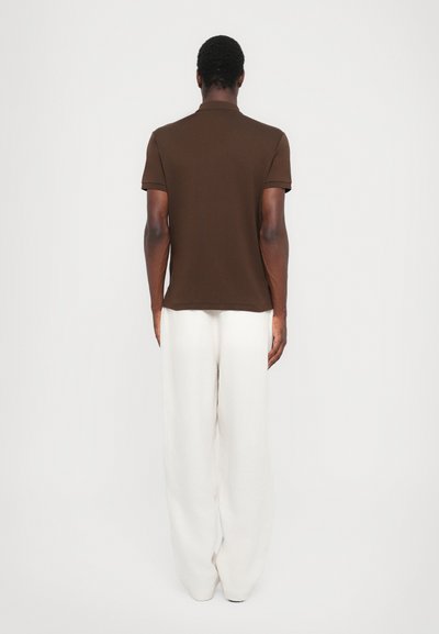 Camisa polo marrón de manga corta, fabricada en algodón. Combinada con pantalones sueltos de color crema. Vista desde la parte de atrás.