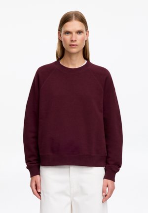 Femme aux cheveux blonds et raides porte un sweat-shirt uni à manches longues rouge foncé et un pantalon blanc, debout devant un fond blanc.