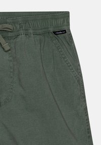 Pantaloni corti verdi con vita elastica, coulisse e tasche laterali; realizzati in tessuto leggero, con cuciture texturizzate e una patch con logo.