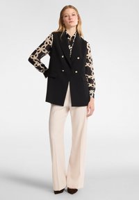 Schwarzer ärmelloser Blazer mit goldenen Knöpfen, kombiniert mit einer gemusterten Bluse in Creme und Schwarz sowie beigen, weit geschnittenen Hosen.