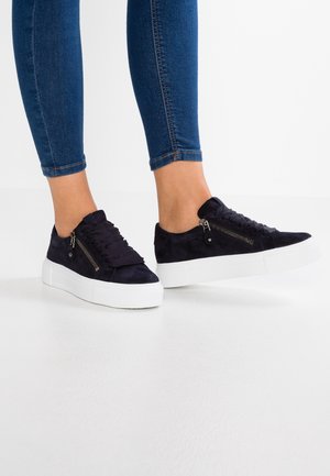 Fødder iført marineblå ruskindssneakers med hvide tykke såler og sideskydelåse, kombineret med tætsiddende blå jeans mod en enkel hvid baggrund.