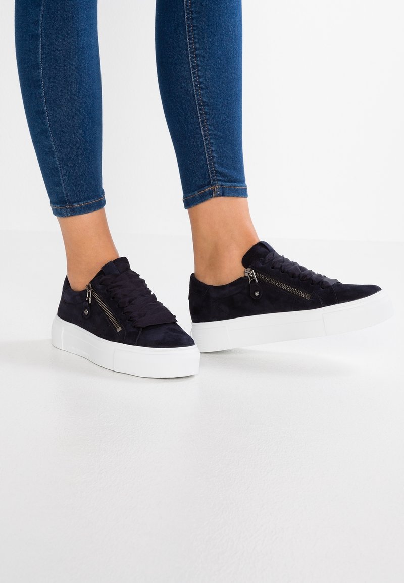 Pieds portant des baskets en daim bleu marine avec des semelles blanches épaisses et des fermetures éclair latérales, assorties à un jean bleu ajusté sur un fond blanc uni.