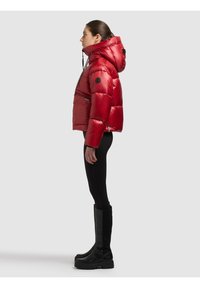 Nainen profiilissa, yllään punainen toppatakki hupulla, mustat leggingsit ja mustat korkeat saappaat, seisomassa yksinkertaisen taustan edessä.