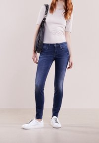Jeans ajustados en denim azul oscuro, con acentos desgastados. Combinados con una camiseta ajustada gris claro y zapatillas blancas. Incluye un bolso negro.