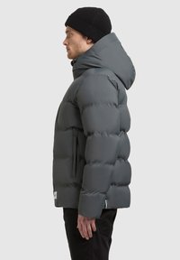 Grijze pufferjack met capuchon, heeft een gewatteerd ontwerp, dikke isolatie en zijzakken. Draagt een zwarte gebreide muts.