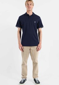 Polo bleu marine avec bordure blanche, en coton, manches courtes, porté avec un pantalon beige et des baskets noires.
