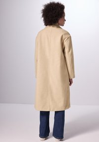 Cappotto di lana beige dal design minimalista, con taglio dritto, colletto a risvolto e maniche lunghe, abbinato a jeans scuri e scarpe bianche.