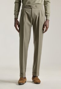 Pantalones verdes oliva con pliegues y un corte ajustado, piernas entalladas y dobladillos rematados. Combinados con mocasines de ante marrón. Textura suave.