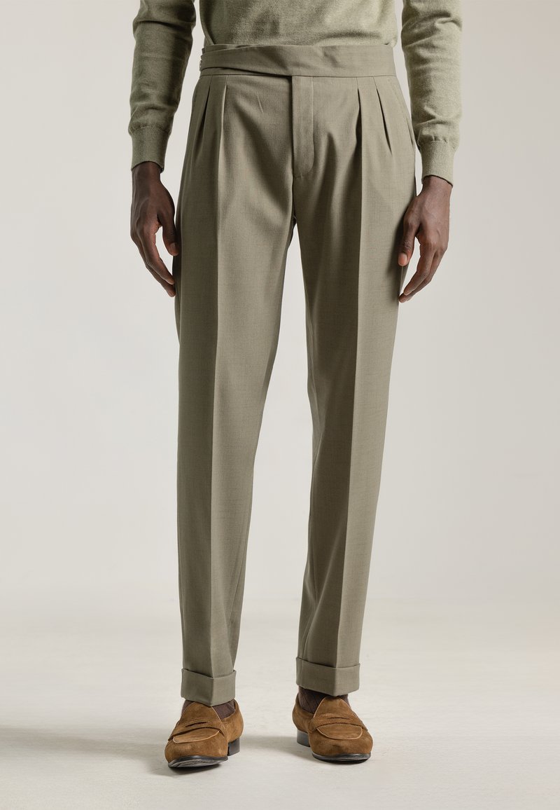 Pantalones verdes oliva con pliegues y un corte ajustado, piernas entalladas y dobladillos rematados. Combinados con mocasines de ante marrón. Textura suave.