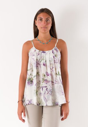Donna che indossa una camicia bianca senza maniche con motivi floreali viola e verdi, pantaloni beige, collana turchese, in piedi davanti a uno sfondo semplice.