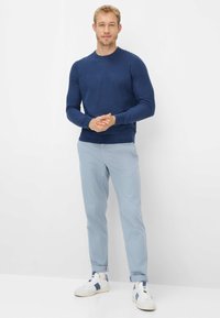 Maglione blu lavorato a maglia, pantaloni azzurri chiari e sneakers bianche con dettagli blu. Modello in piedi con le mani incrociate su uno sfondo bianco.
