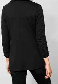 Person som bär en figursydd svart kavaj med rynkiga ärmar, sedd från baksidan mot en enkel ljusgrå bakgrund.