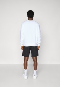 Weißer Pullover mit langen Ärmeln, kombiniert mit schwarzen Shorts. Modell trägt weiße Sneakers und knöchellange weiße Socken. Einfaches Design.