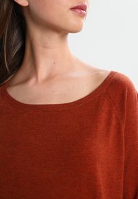 Jeune femme portant un pull en maille couleur rouille avec un large décolleté, montrant le cou, l'épaule, ainsi qu'une partie de ses lèvres et de ses cheveux.