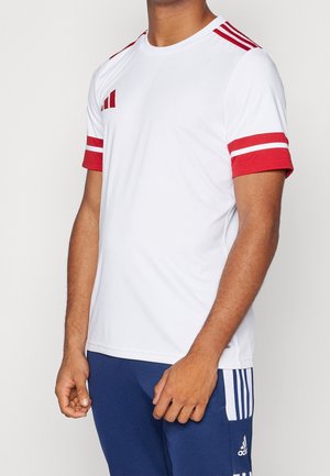 Mężczyzna w białej koszulce z czerwonymi paskami na ramionach i rękawach, w połączeniu z niebieskimi spodniami Adidas z białymi paskami.