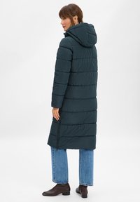 Turkusowa kurtka puchowa z kapturem, z pikowanym wzorem, długim krojem i bocznym zamkiem. Nosi się ją z niebieskimi spodniami typu wide-leg i brązowymi botkami za kostkę.