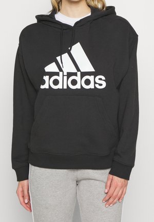Vrouw draagt een zwarte Adidas-hoodie met een wit logo en lichtgrijze joggingbroek, staat met ontspannen handen langs haar zij.