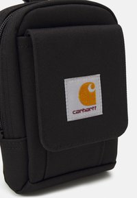 Saco de tecido preto com aba frontal, apresentando um rótulo branco com um logótipo amarelo e "carhartt" impresso em vermelho. Fecho com zíper.
